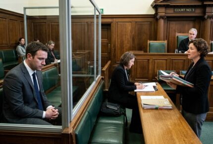 A man in a grey suit and blue tie sits in the glass dock of UK Court No. 3 looking downcast while his female barrister presents bail application case files to the judge, with a solicitor taking notes nearby and members of the public seated in the gallery behind the dock.