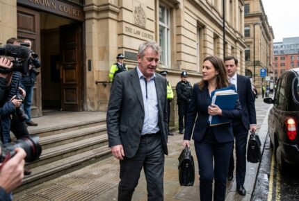 A middle-aged man in a grey suit walks away from a UK Crown Court alongside his solicitor carrying blue legal folders and a briefcase, with press photographers filming, police officers standing guard at the entrance, and a black taxi waiting on the street.
