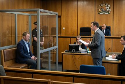 A man in a navy blazer sits in the glass dock of a UK Crown Court during a bank fraud bail hearing while his barrister in a grey suit reads from case papers, with the judge presiding under the Royal Coat of Arms, a court clerk typing on a computer, a uniformed police officer guarding the dock, and another barrister taking notes.
