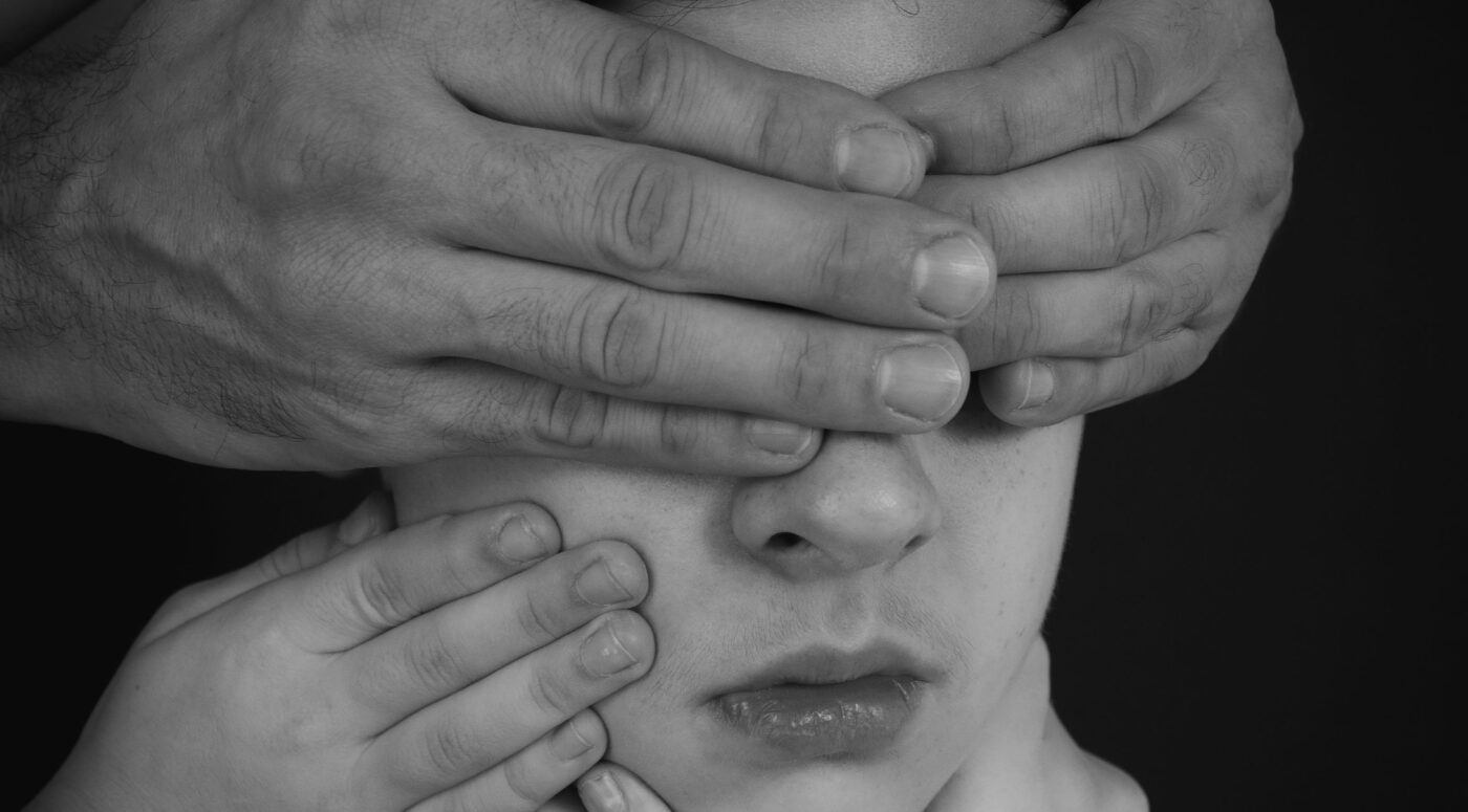 A black-and-white portrait of a young person whose face is obscured by multiple hands reaching from different directions — large hands cover their eyes and forehead while smaller hands press against their cheeks and chin, creating a striking, unsettling composition against a dark background that evokes themes of silencing, control, or overwhelming external pressure.