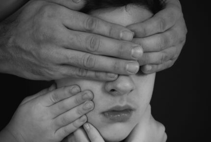 A black-and-white portrait of a young person whose face is obscured by multiple hands reaching from different directions — large hands cover their eyes and forehead while smaller hands press against their cheeks and chin, creating a striking, unsettling composition against a dark background that evokes themes of silencing, control, or overwhelming external pressure.